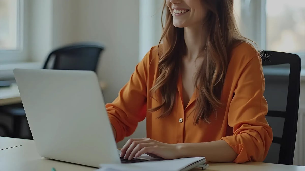 Mujer sonriente de camisa naranja trabajando en una laptop en una oficina luminosa, con una libreta sobre el escritorio.