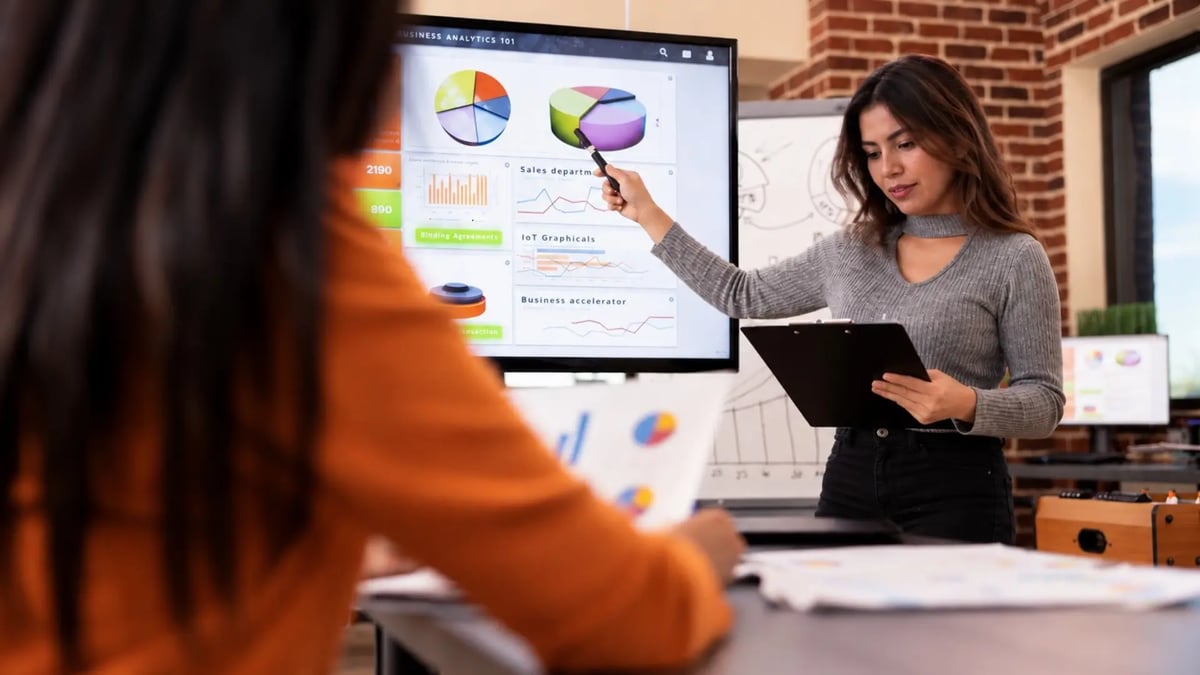 Una profesional de pollera y remera gris señala con un marcador una pantalla con un dashboard de analítica de negocio —gráficos de torta, barras y líneas— mientras sostiene una tablet; en primer plano, de espaldas, una colega de camisa naranja la escucha durante una reunión de trabajo.